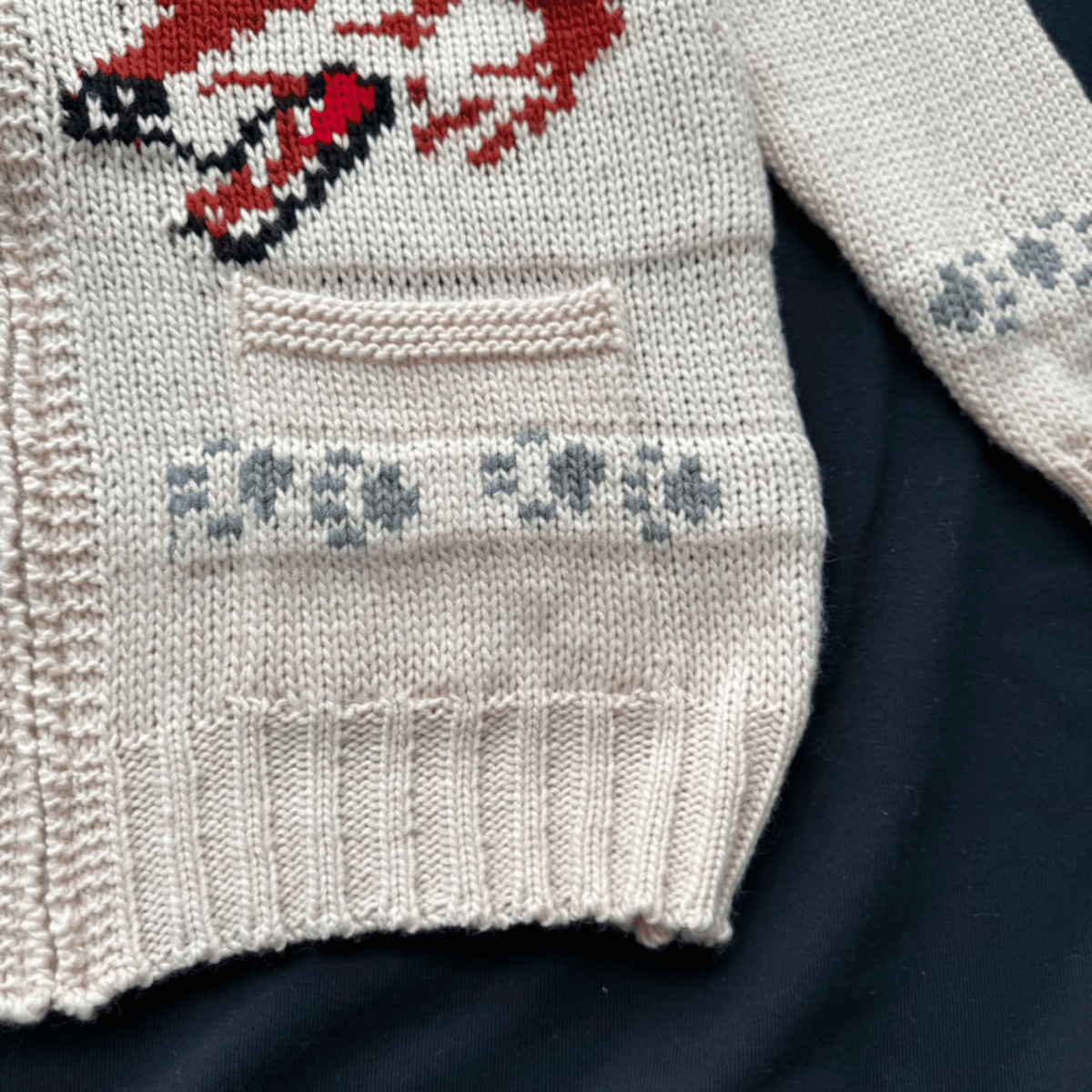 Close-up of a knitted sweater in cream with a red and black graphic motif and gray snowflake patterns, laid flat on a dark navy background.