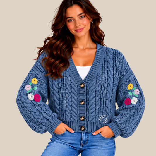 A woman wearing a blue cable-knit cardigan with colorful floral embroidery on both sleeves, styled with blue jeans and a white top, set against a warm beige background.