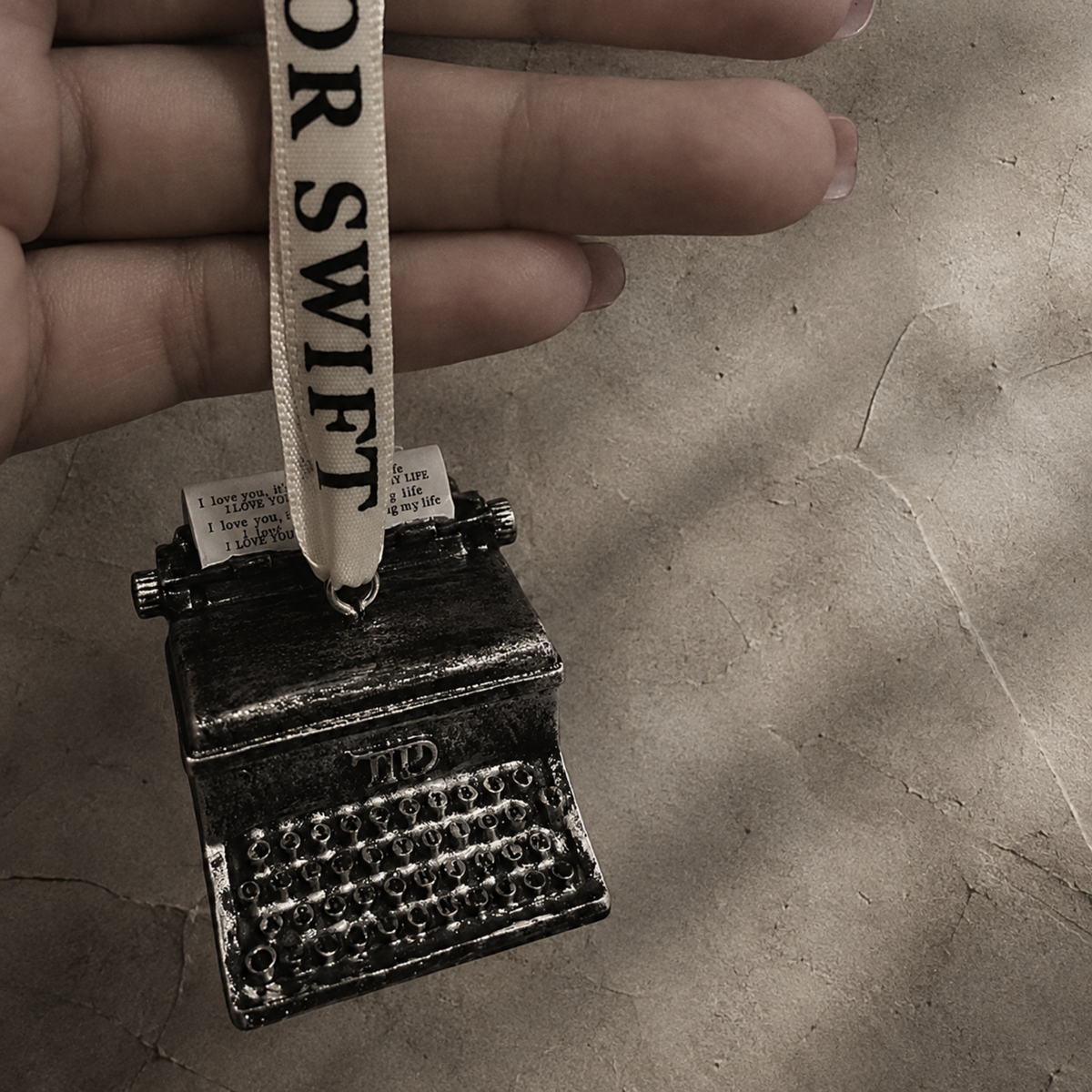 A miniature typewriter ornament held in a hand, featuring a dark metallic body with a cream ribbon hanger printed with the name "Swift"