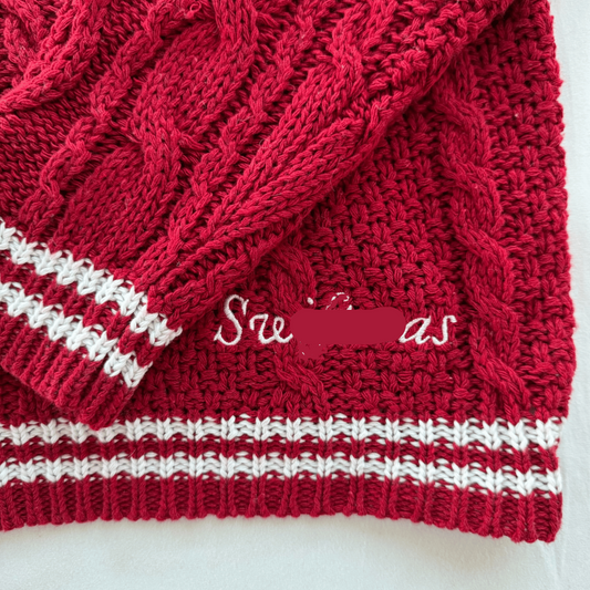 A close-up of a red cable-knit sweater or scarf with white striped ribbed trim and white cursive text reading 'Swiftmas' across the front, photographed on a light background.