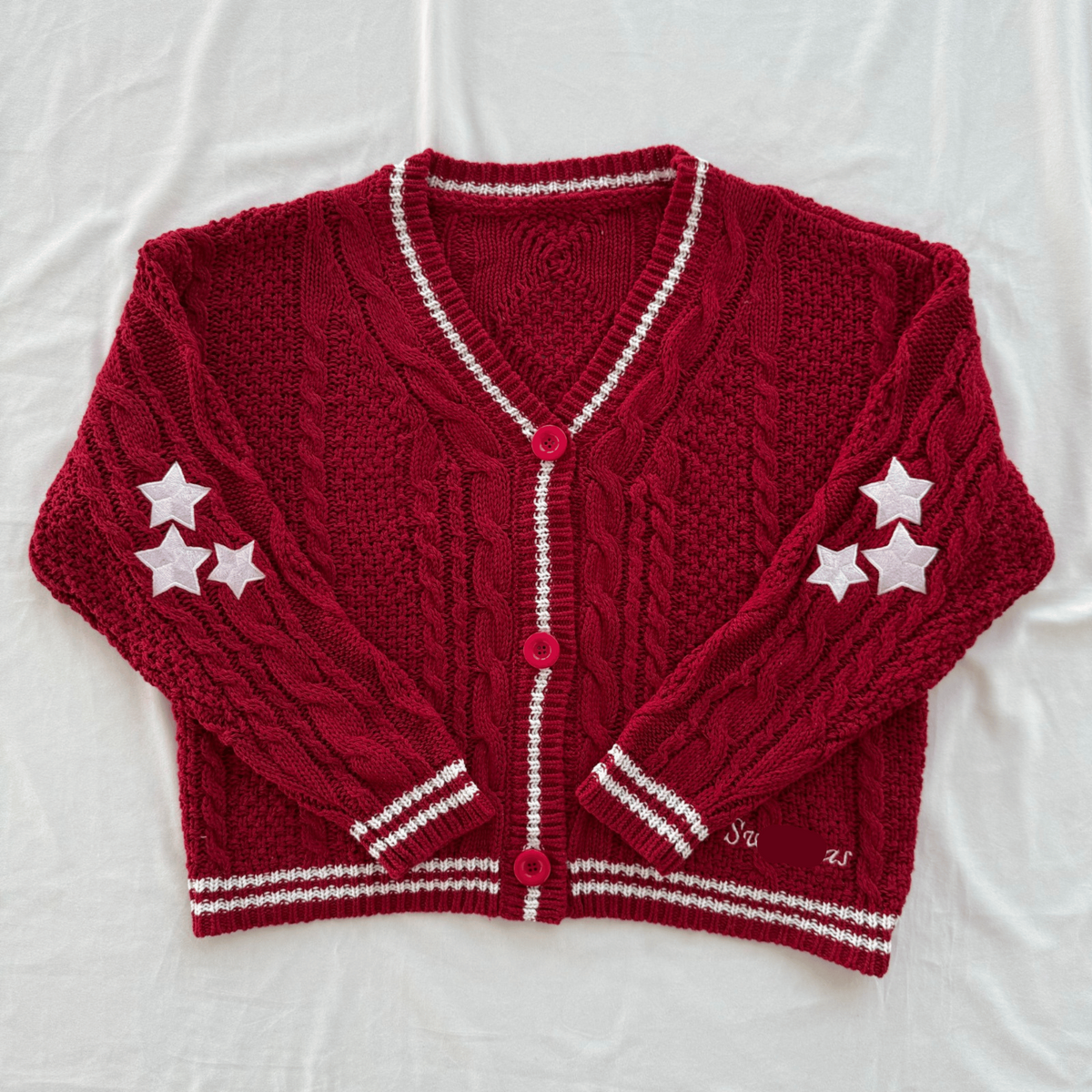 A red cable-knit cardigan sweater with white trim, featuring three white stars on each sleeve and a white embroidered 'Swiftmas' brand name on the lower right front. The cardigan is laid flat on a white fabric background.