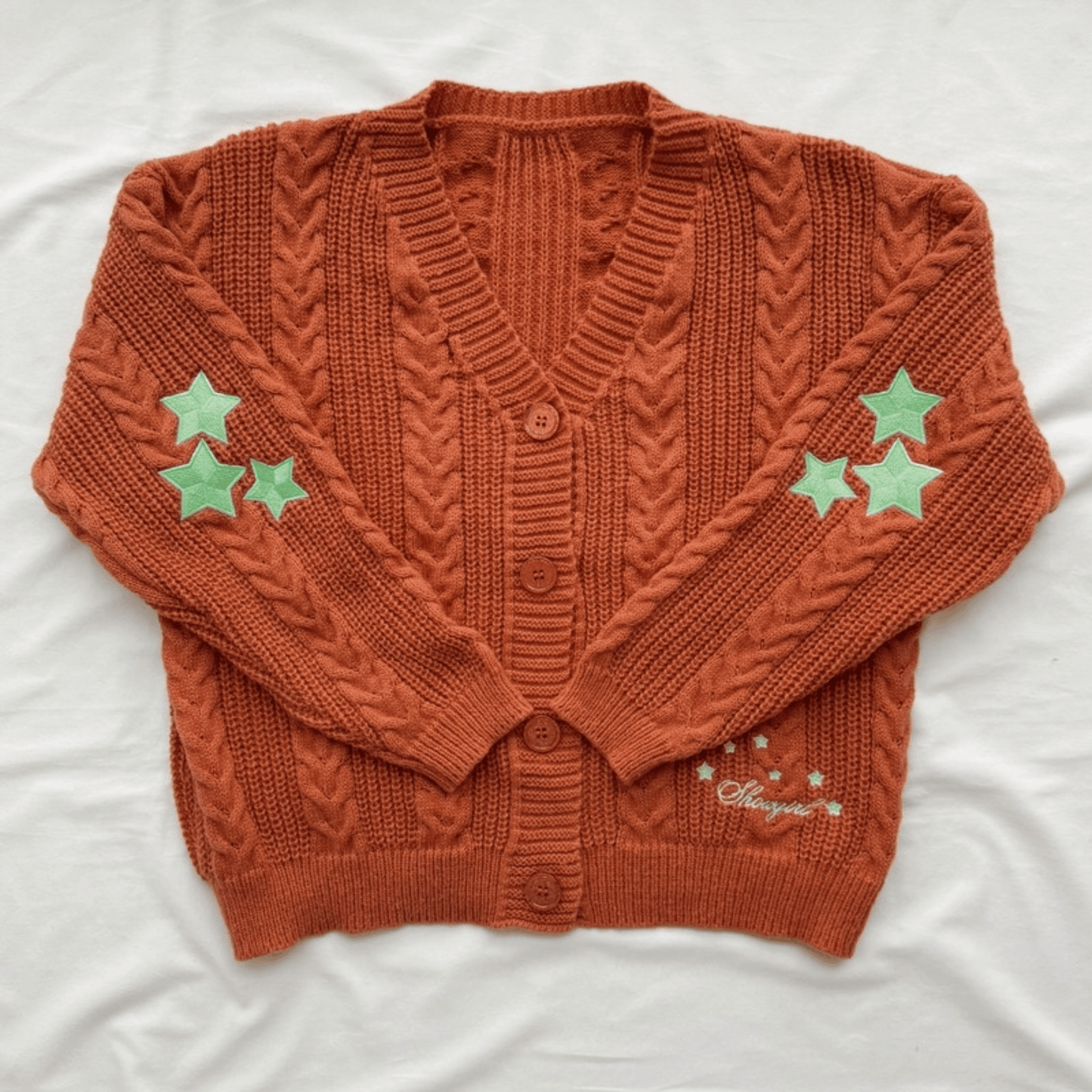 An orange cable-knit cardigan sweater with three light green star patches on each sleeve and a small embroidered script on the lower right front, laid flat on a white fabric background.