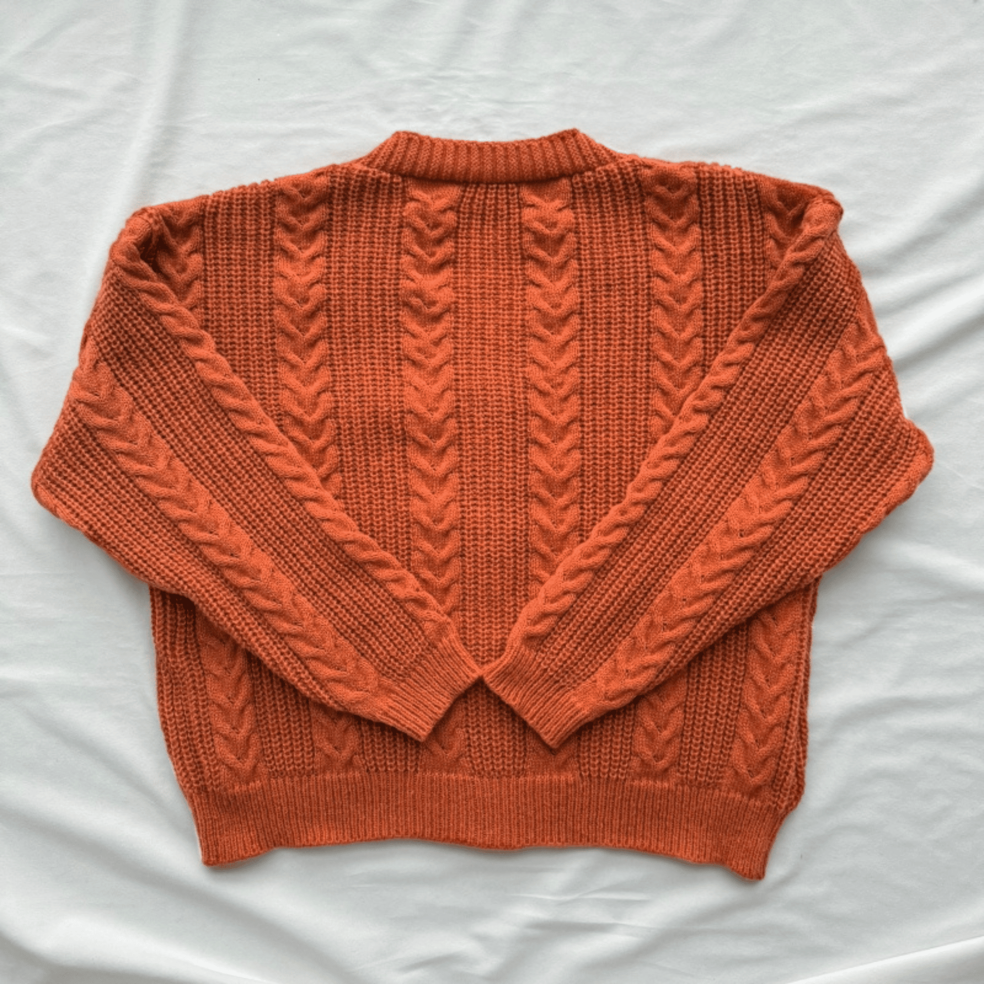 An orange cable-knit sweater laid flat on a white fabric background.