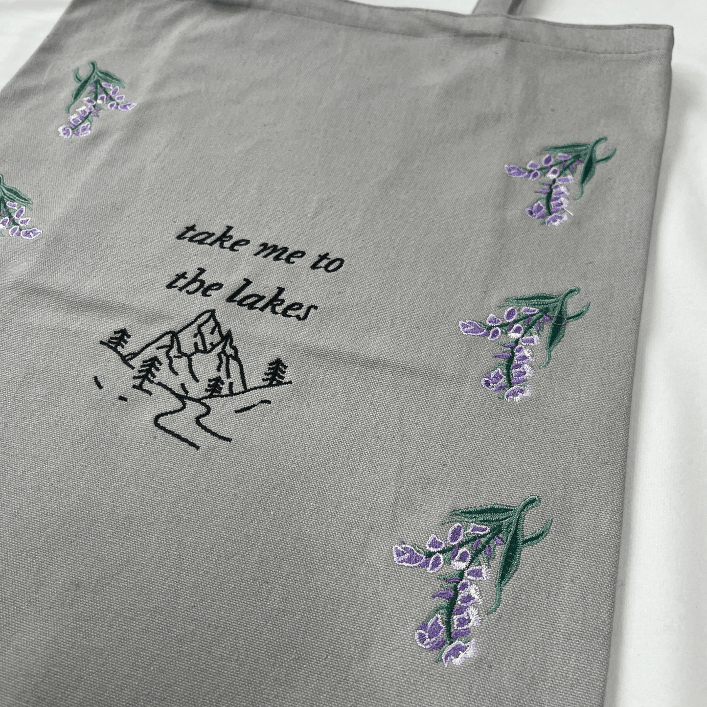 Gray fabric with embroidered lavender flowers and 'take me to the lakes' text on a white background