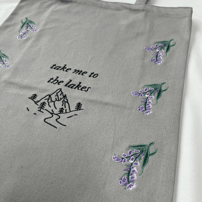 Gray fabric with embroidered lavender flowers and 'take me to the lakes' text on a white background