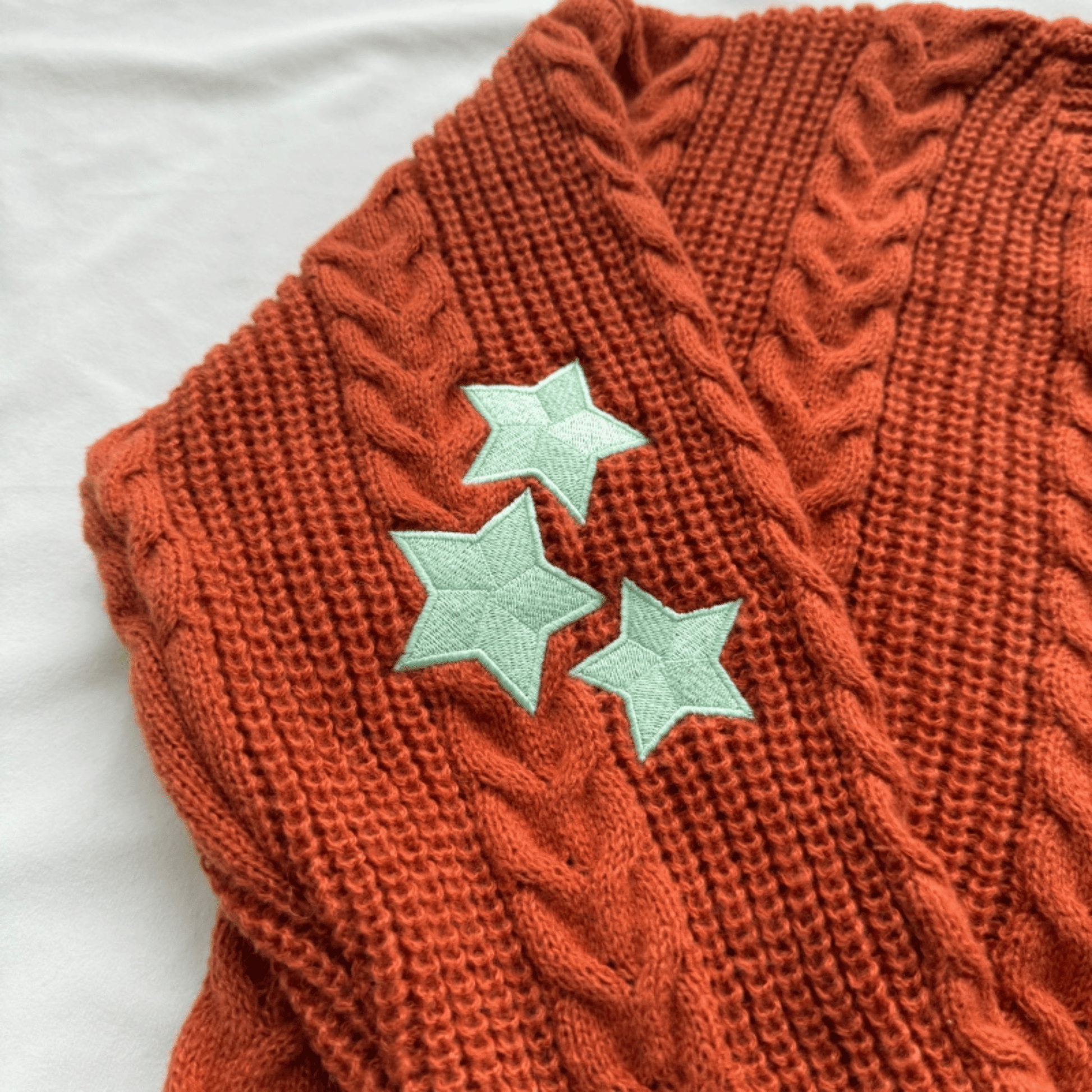 A close-up of a rust-orange cable-knit sweater vest featuring three mint green embroidered star appliqués on the front, laid flat on a white fabric background.
