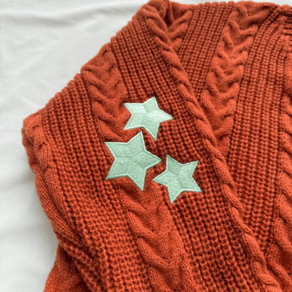 A close-up of a rust-orange cable-knit sweater vest featuring three mint green embroidered star appliqués on the front, laid flat on a white fabric background.