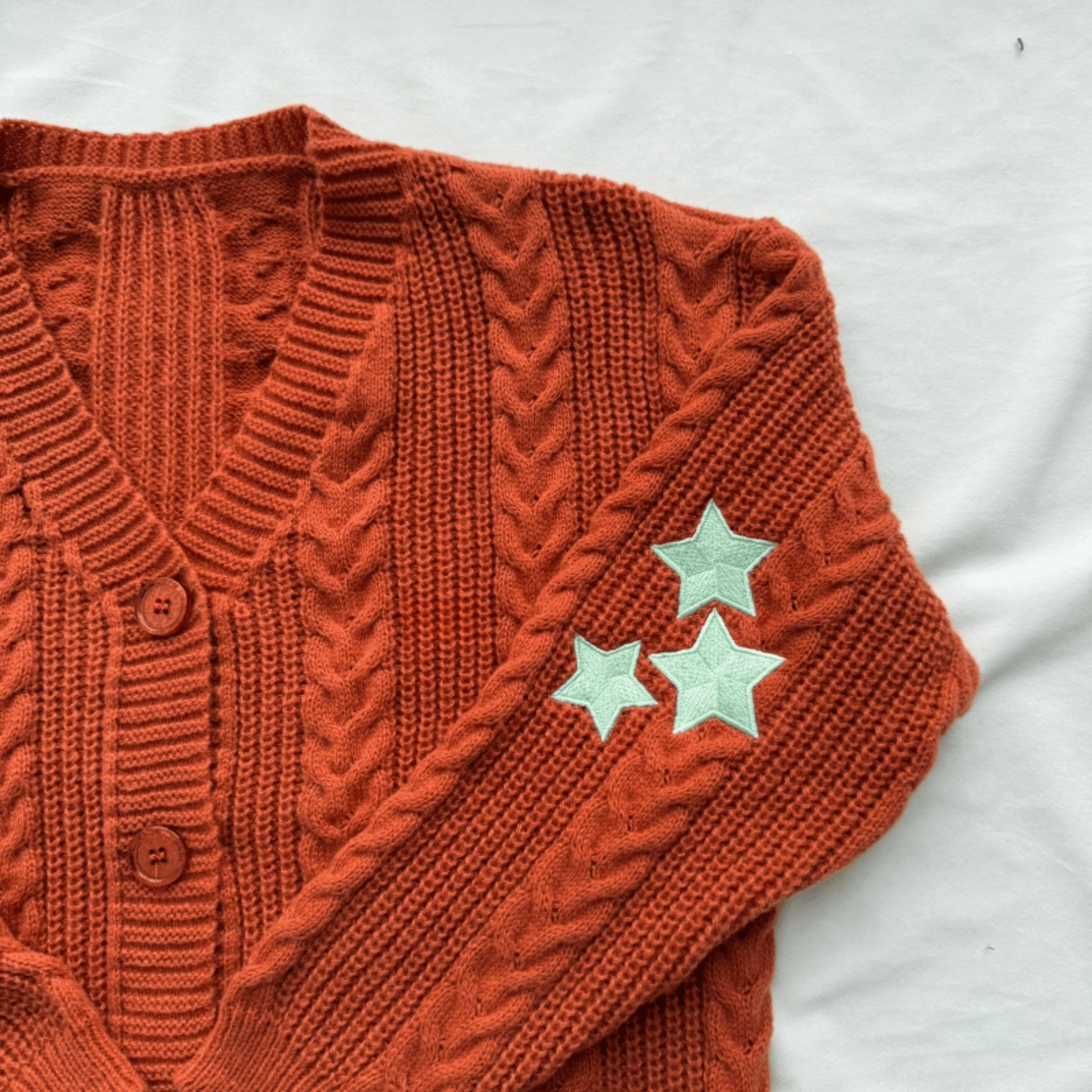 A close-up of a rust-orange cable-knit cardigan sweater with three mint green embroidered stars on the sleeve, laid flat on a white fabric background.