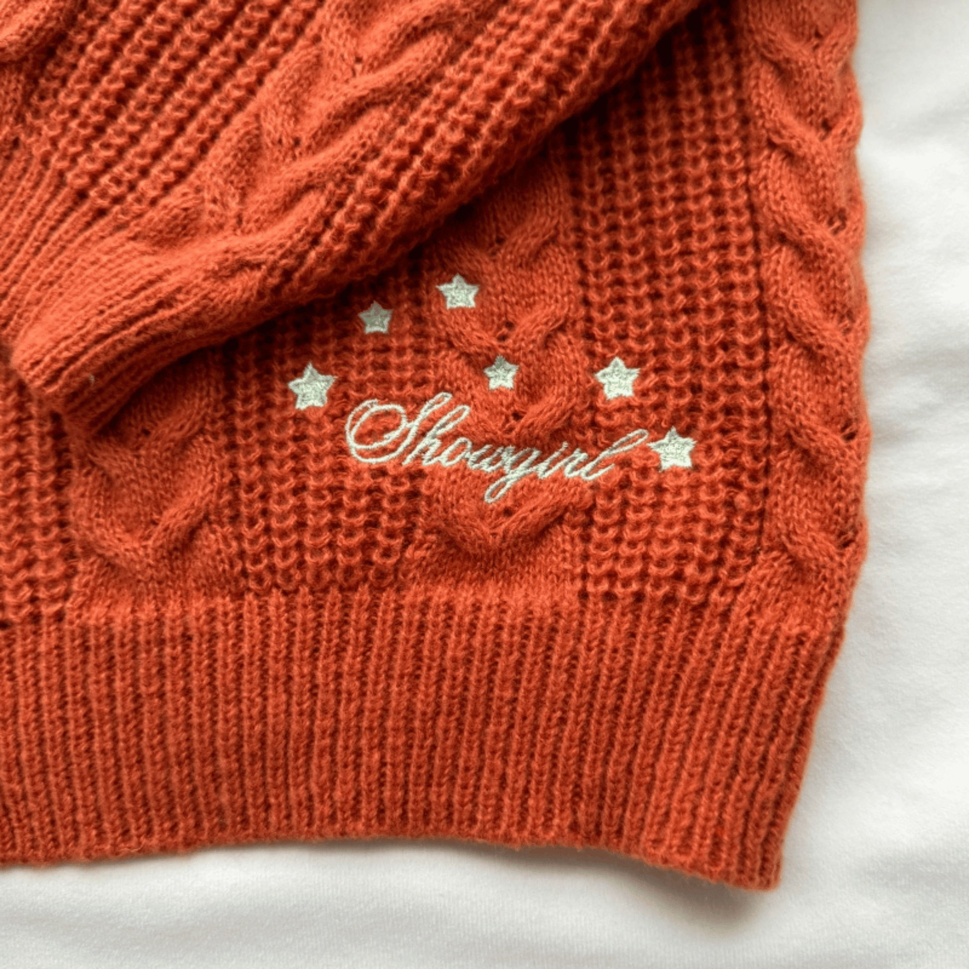 A close-up of a textured orange cable-knit sweater with white embroidered script reading 'Showgirl' and small white stars, laid flat on a white fabric background.