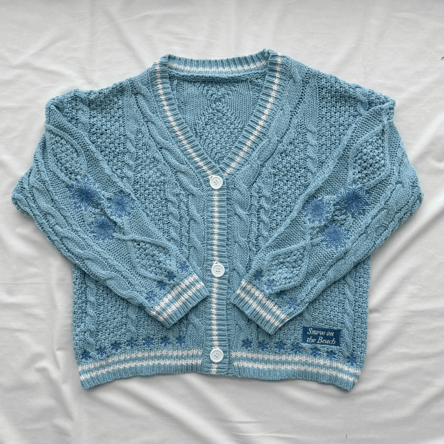 A light blue cable-knit cardigan sweater with white striped trim and white buttons, laid flat on a white fabric background. A small woven label on the lower right reads 'Snow On The Beach'.
