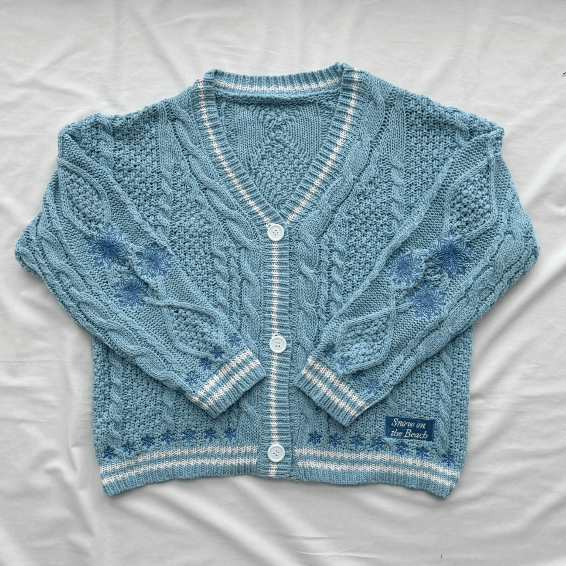 A light blue cable-knit cardigan sweater with white striped trim and white buttons, laid flat on a white fabric background. A small woven label on the lower right reads 'Snow On The Beach'.