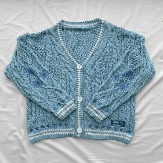 A light blue cable-knit cardigan sweater with white striped trim and white buttons, laid flat on a white fabric background. A small woven label on the lower right reads 'Snow On The Beach'.