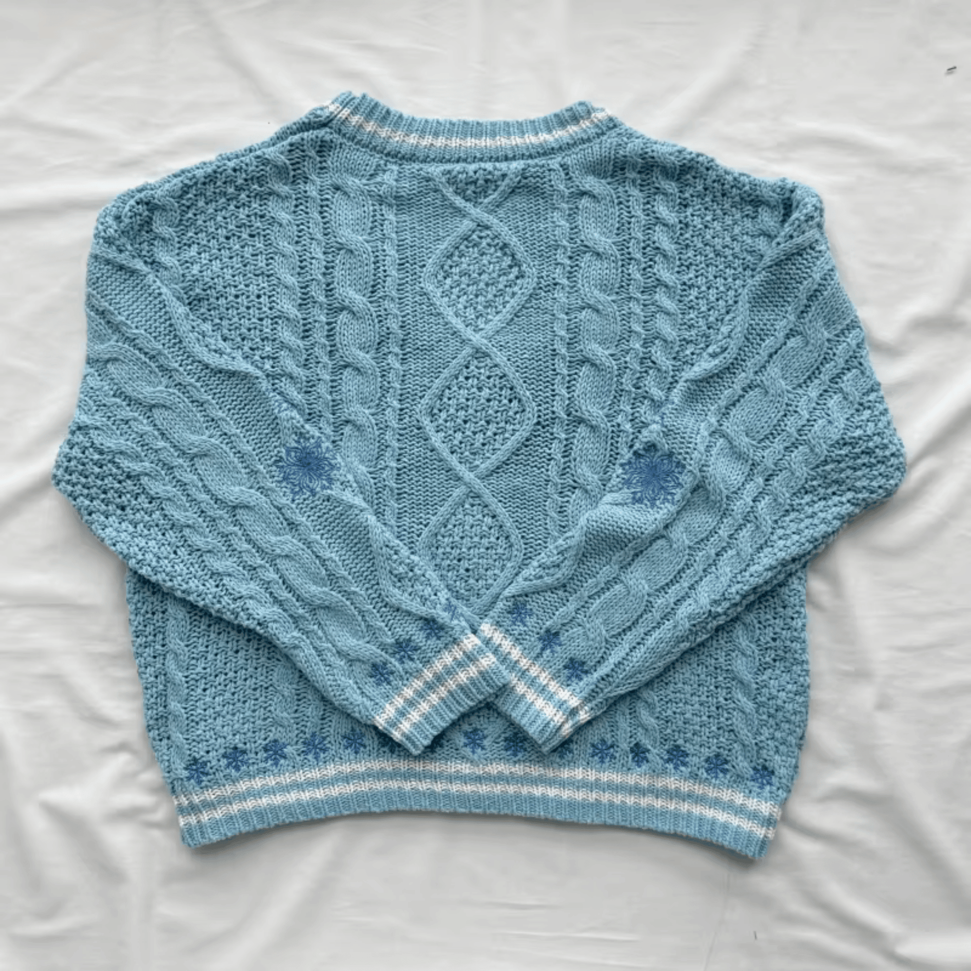 A light blue cable-knit sweater laid flat on a white fabric background, showing the back design with diamond and cable patterns and striped ribbed cuffs and hem.