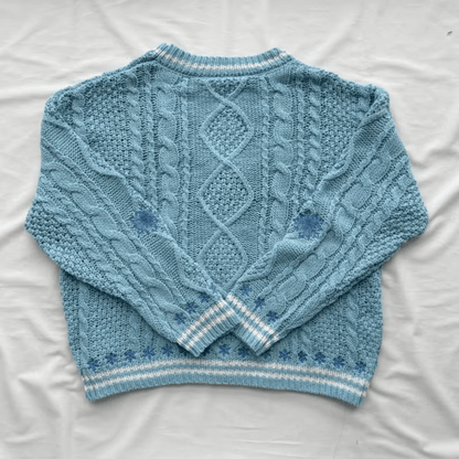 A light blue cable-knit sweater laid flat on a white fabric background, showing the back design with diamond and cable patterns and striped ribbed cuffs and hem.