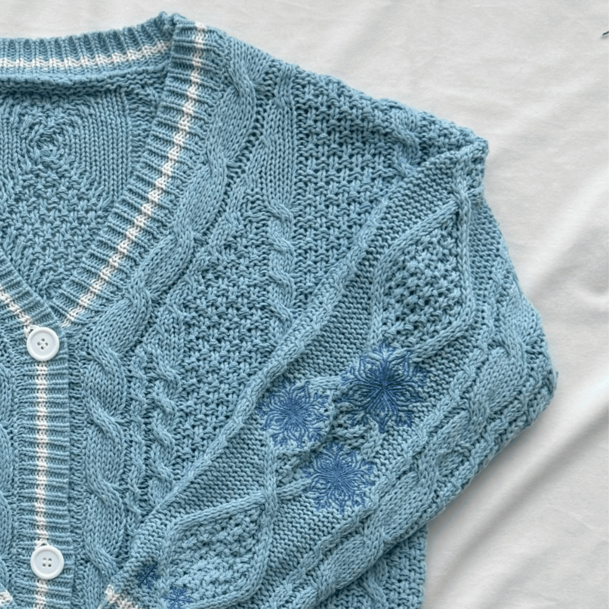 A light blue cable-knit cardigan sweater with white buttons, laid flat on a white fabric background.