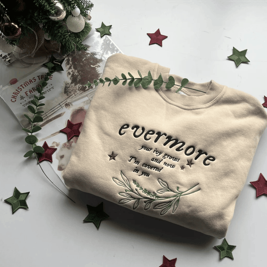 Beige sweatshirt with text and greenery on a white surface with decorative stars and a Christmas tree.