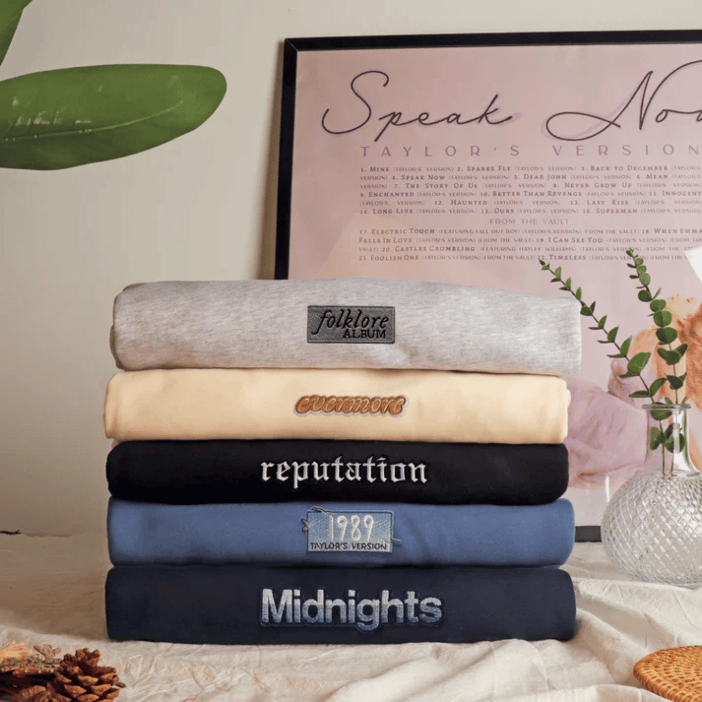 A stack of five folded sweatshirts in different colors, each with a custom embroidered or printed label, displayed on a tablecloth with decorative props like a framed print and a vase.