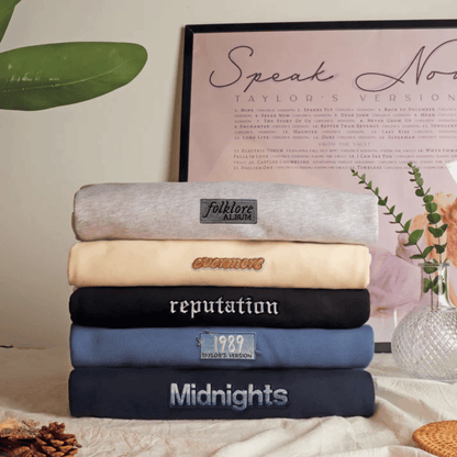 A stack of five folded sweatshirts in different colors, each with a custom embroidered or printed label, displayed on a tablecloth with decorative props like a framed print and a vase.