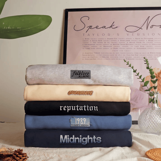 A stack of five folded sweatshirts in different colors, each with a custom embroidered or printed label, displayed on a tablecloth with decorative props like a framed print and a vase.