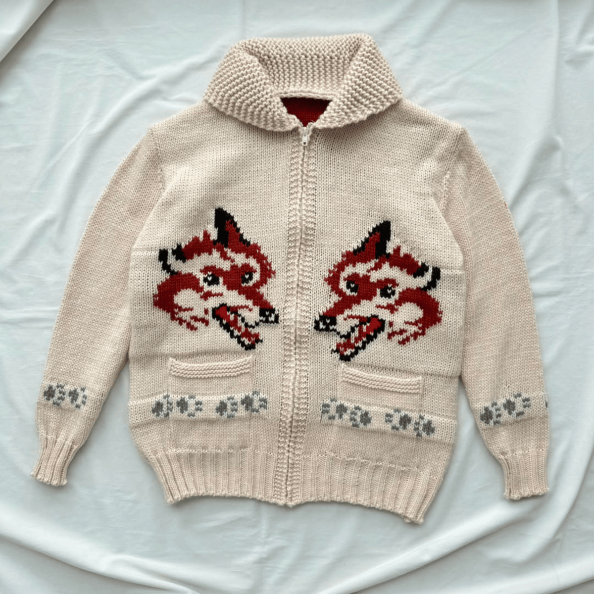 A cream-colored knit zip-up cardigan sweater featuring two large red and black fox faces on the front, with gray paw-print patterns near the lower front pockets. The cardigan is laid flat on a white fabric background.
