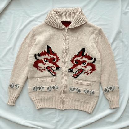 A cream-colored knit zip-up cardigan sweater featuring two large red and black fox faces on the front, with gray paw-print patterns near the lower front pockets. The cardigan is laid flat on a white fabric background.