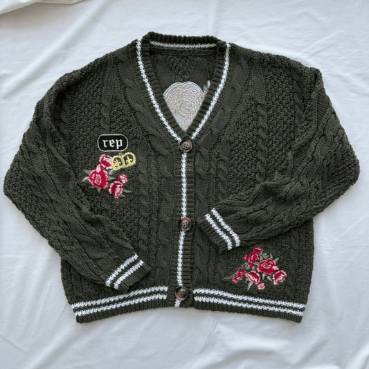 An olive green cable-knit cardigan sweater with white striped trim, brown buttons, and pink rose embroidery, laid flat on a white fabric background.