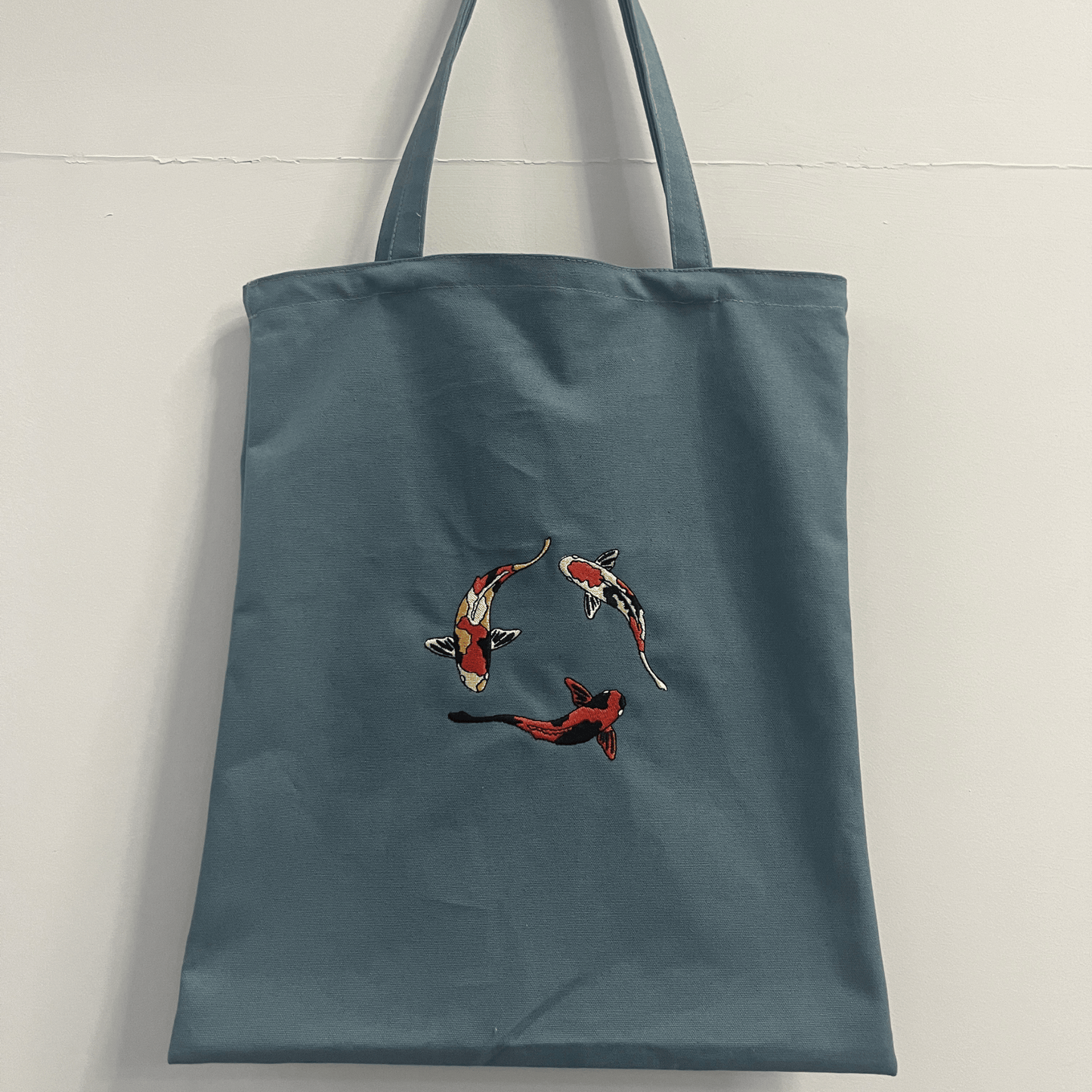 Blue tote bag with embroidered koi fish design on a light gray background