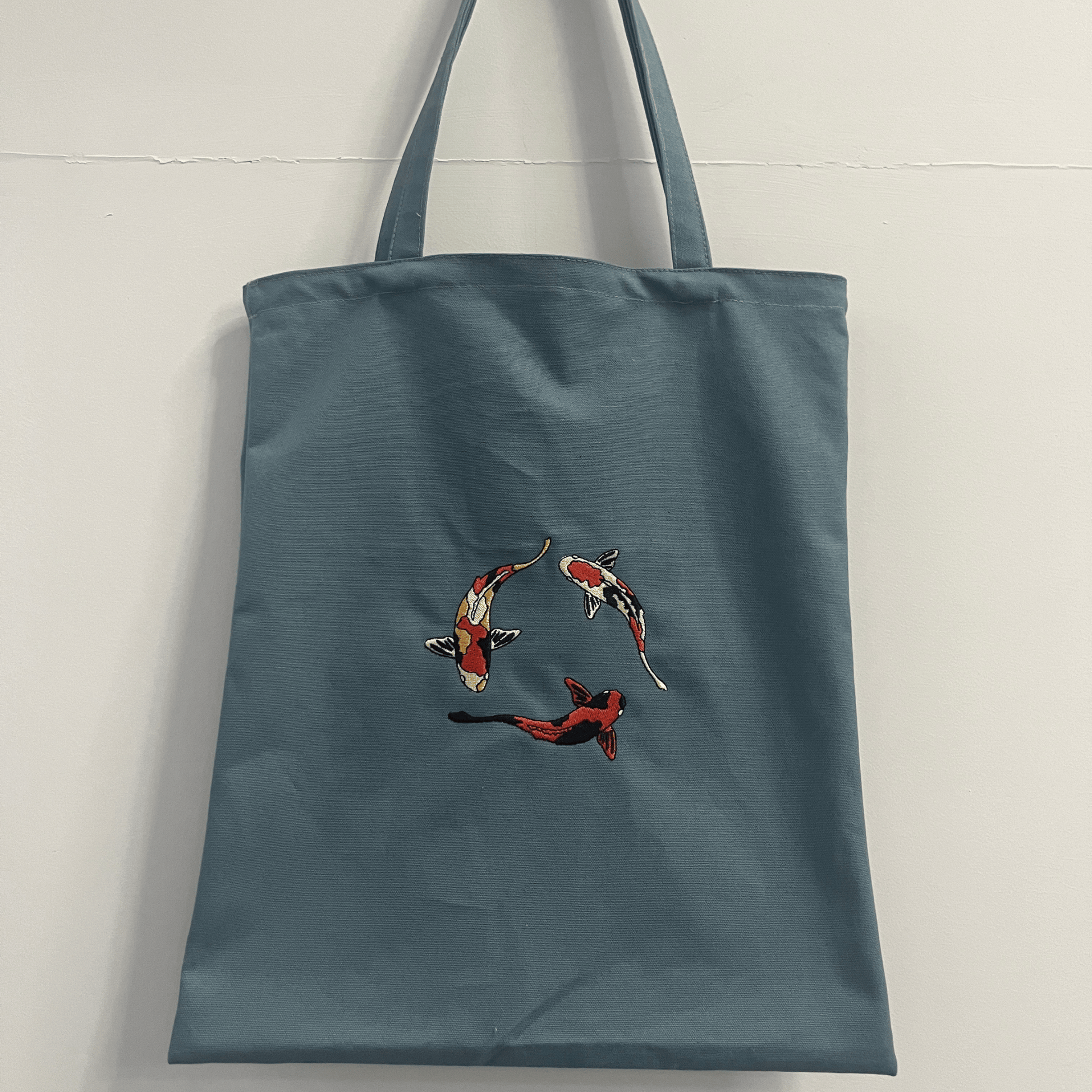 Blue tote bag with embroidered koi fish design on a light gray background