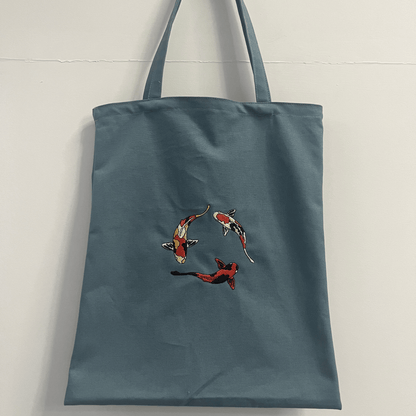 Blue tote bag with embroidered koi fish design on a light gray background