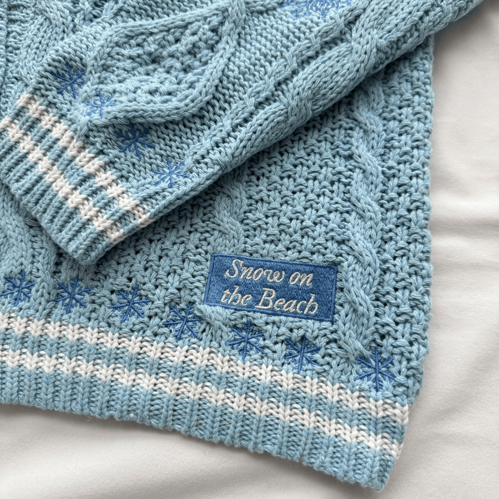 A close-up of a light blue cable-knit sweater with white stripes, embroidered blue snowflakes, and a blue label reading 'Snow on the Beach'.