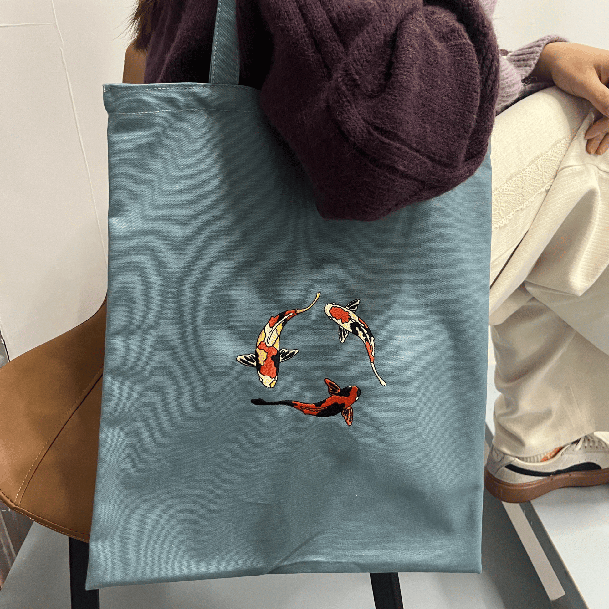 Blue tote bag with koi fish design held by a person sitting on a chair.