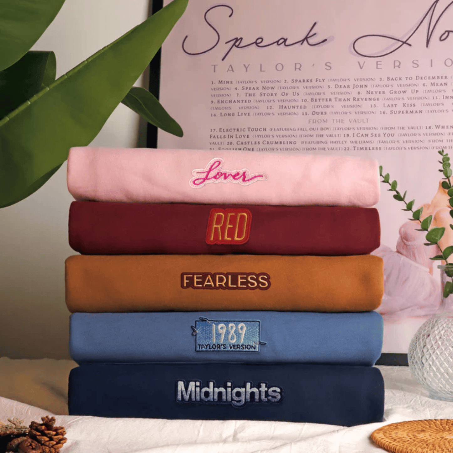 A stack of six folded sweatpants in different colors, each with a unique graphic text design, displayed on a table with decorative props including a green leaf, a framed print, and a woven basket.