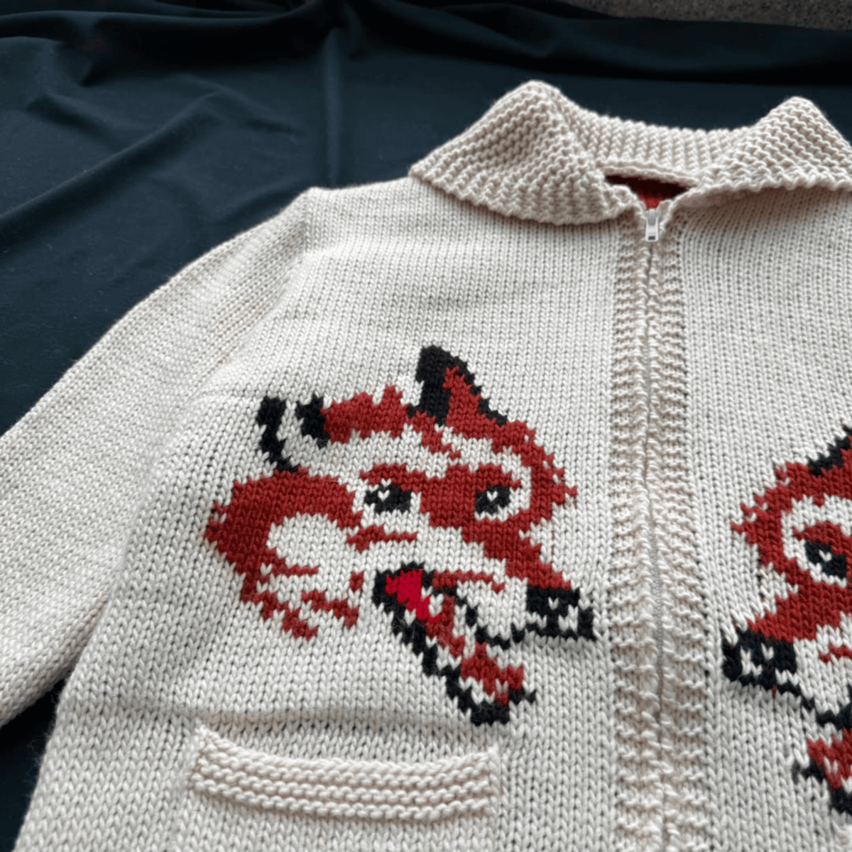 A close-up of a cream-colored hand-knit zip-up sweater featuring a red, black, and white fox face motif on the front.