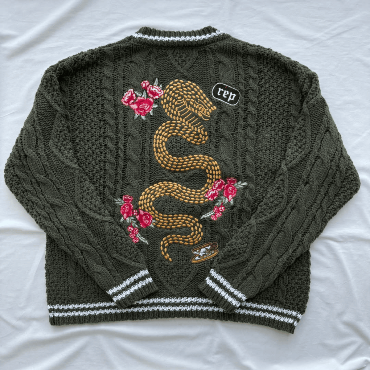 An olive green cable-knit sweater with a large embroidered golden snake and pink roses on the back, finished with white and gray striped ribbed trim, laid flat on a white fabric background.
