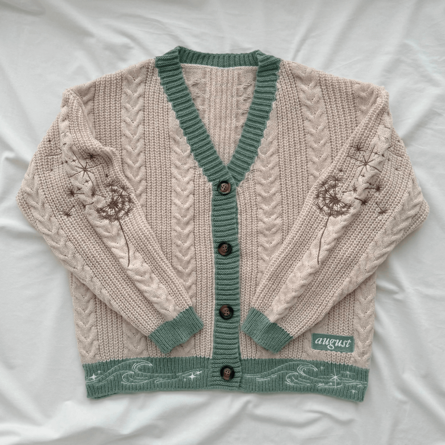 A cream-colored cable-knit cardigan sweater with sage green trim, button closure, and embroidered floral details on the sleeves, laid flat on a white fabric background.