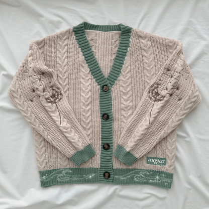 A cream-colored cable-knit cardigan sweater with sage green trim, button closure, and embroidered floral details on the sleeves, laid flat on a white fabric background.