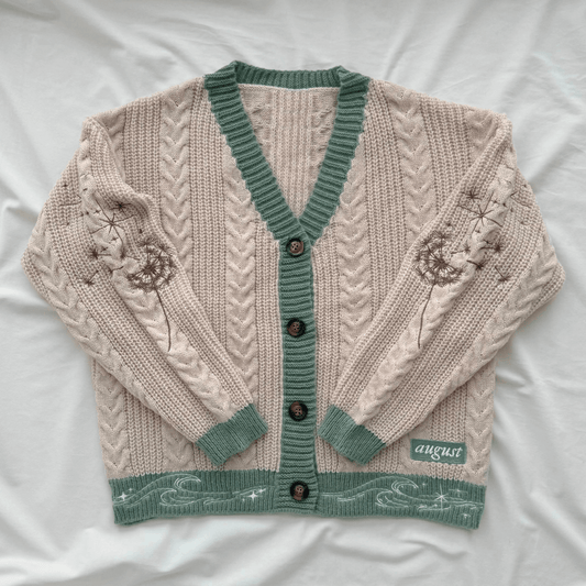 A cream-colored cable-knit cardigan sweater with sage green trim, button closure, and embroidered floral details on the sleeves, laid flat on a white fabric background.