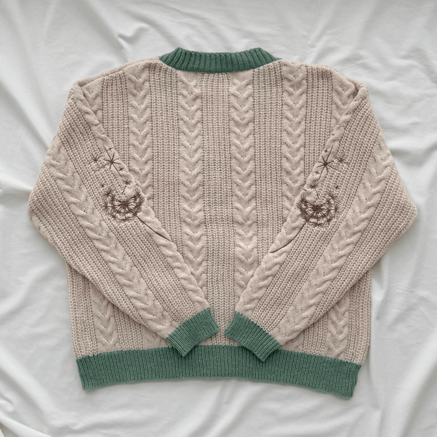 A cream-colored cable-knit sweater with sage green ribbed trim, laid flat on a white fabric background. The back of the sweater features vertical cable knit patterns and embroidered jack-o'-lantern motifs on the upper sleeves.