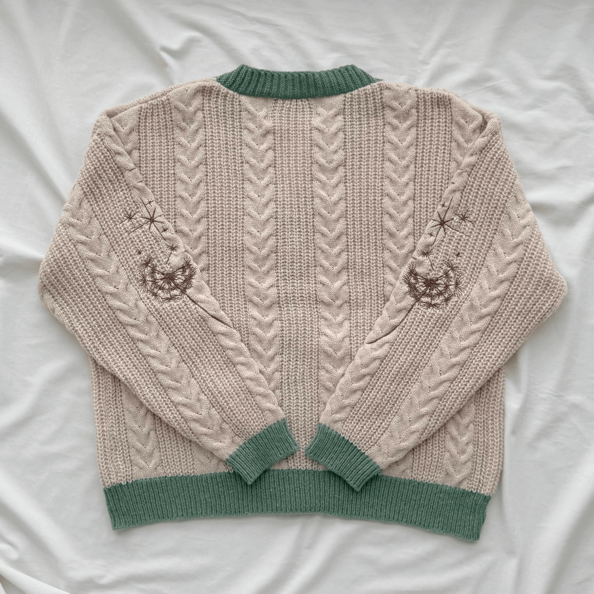 A cream-colored cable-knit sweater with sage green ribbed trim, laid flat on a white fabric background. The back of the sweater features vertical cable knit patterns and embroidered jack-o'-lantern motifs on the upper sleeves.