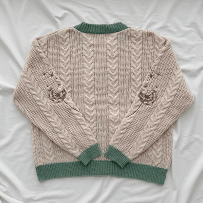 A cream-colored cable-knit sweater with sage green ribbed trim, laid flat on a white fabric background. The back of the sweater features vertical cable knit patterns and embroidered jack-o'-lantern motifs on the upper sleeves.