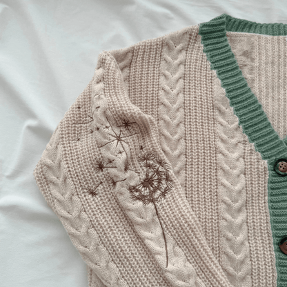 A close-up of a cream-colored cable-knit cardigan with a sage green ribbed trim and brown embroidered dandelion details, laid flat on a white fabric background.