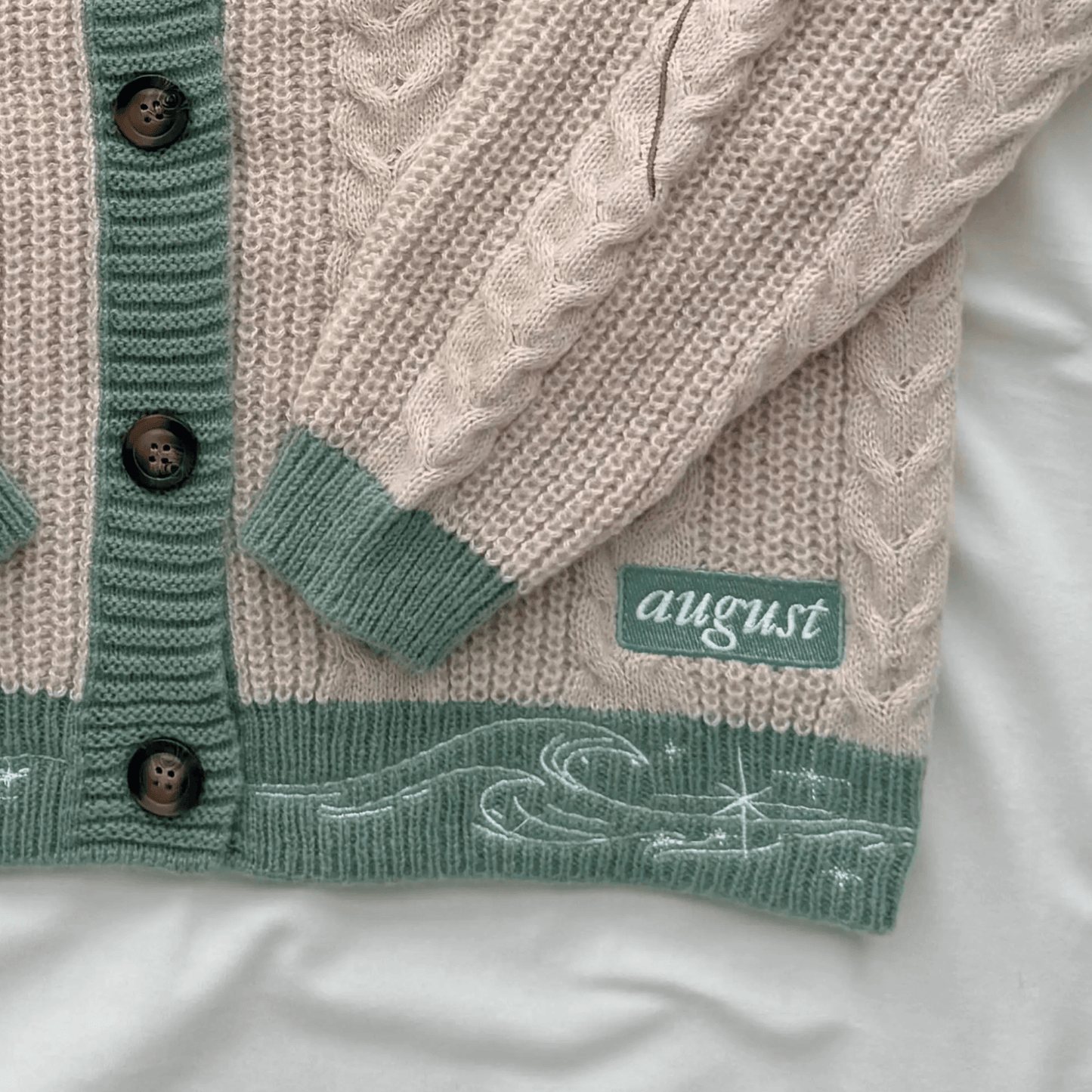 A close-up of a knitted cardigan laid flat on a white fabric background. The cardigan is cream with sage green ribbed trim, dark brown buttons, and a green patch with the word 'august' on it.