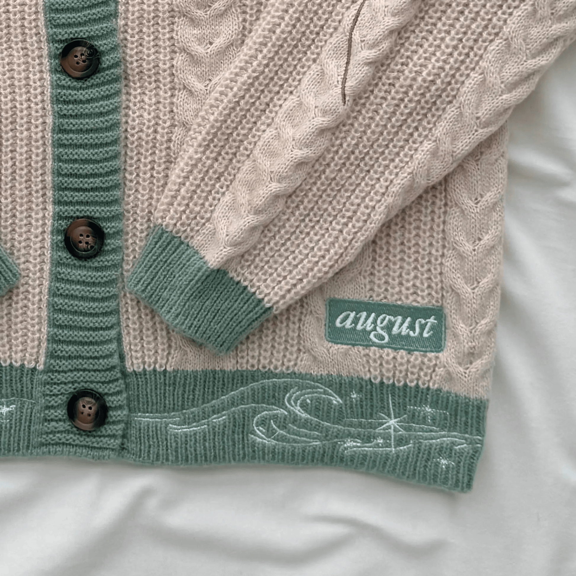 A close-up of a knitted cardigan laid flat on a white fabric background. The cardigan is cream with sage green ribbed trim, dark brown buttons, and a green patch with the word 'august' on it.