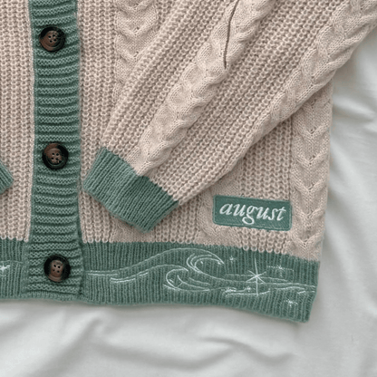 A close-up of a knitted cardigan laid flat on a white fabric background. The cardigan is cream with sage green ribbed trim, dark brown buttons, and a green patch with the word 'august' on it.