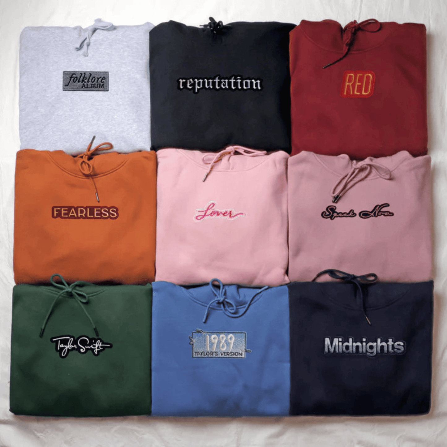 A flat-lay grid of nine folded hoodies in different colors, each with a drawstring hood and a stylized text graphic on the chest. The colors include light gray, black, burgundy, orange, pink, dusty pink, dark green, blue, and navy.