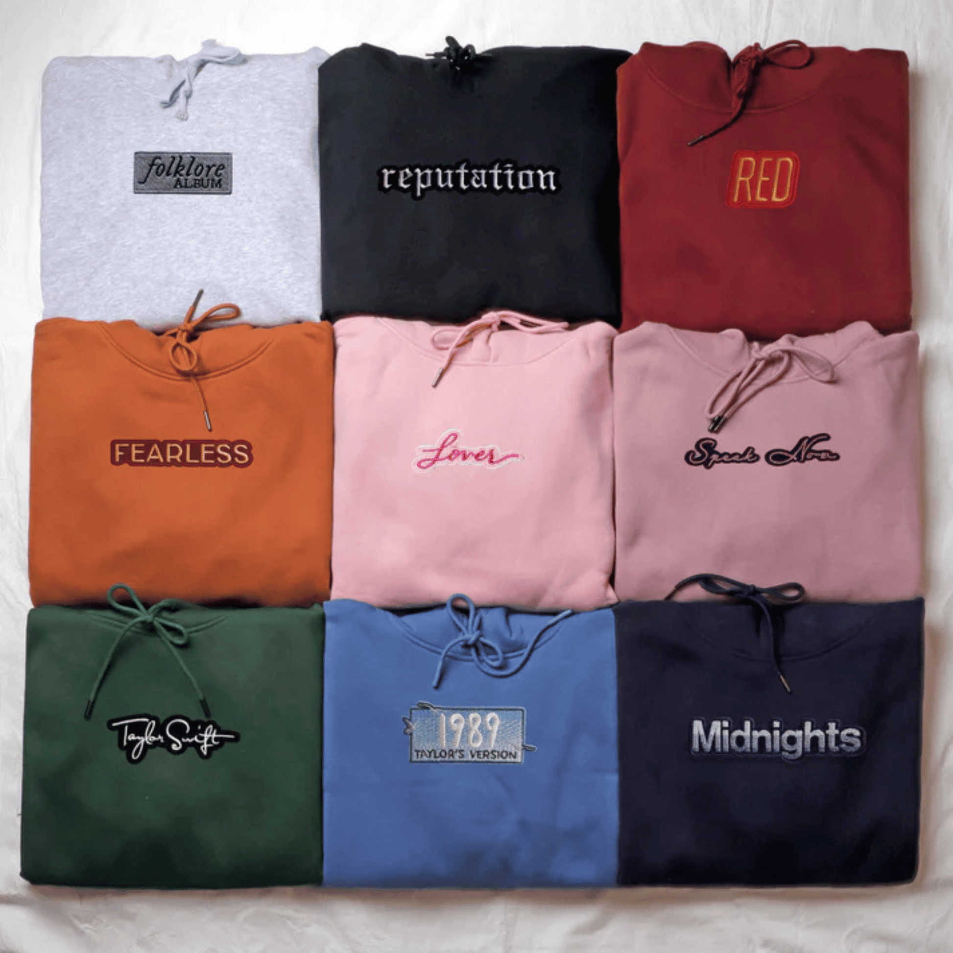 A flat-lay grid of nine folded hoodies in different colors, each with a drawstring hood and a stylized text graphic on the chest. The colors include light gray, black, burgundy, orange, pink, dusty pink, dark green, blue, and navy.