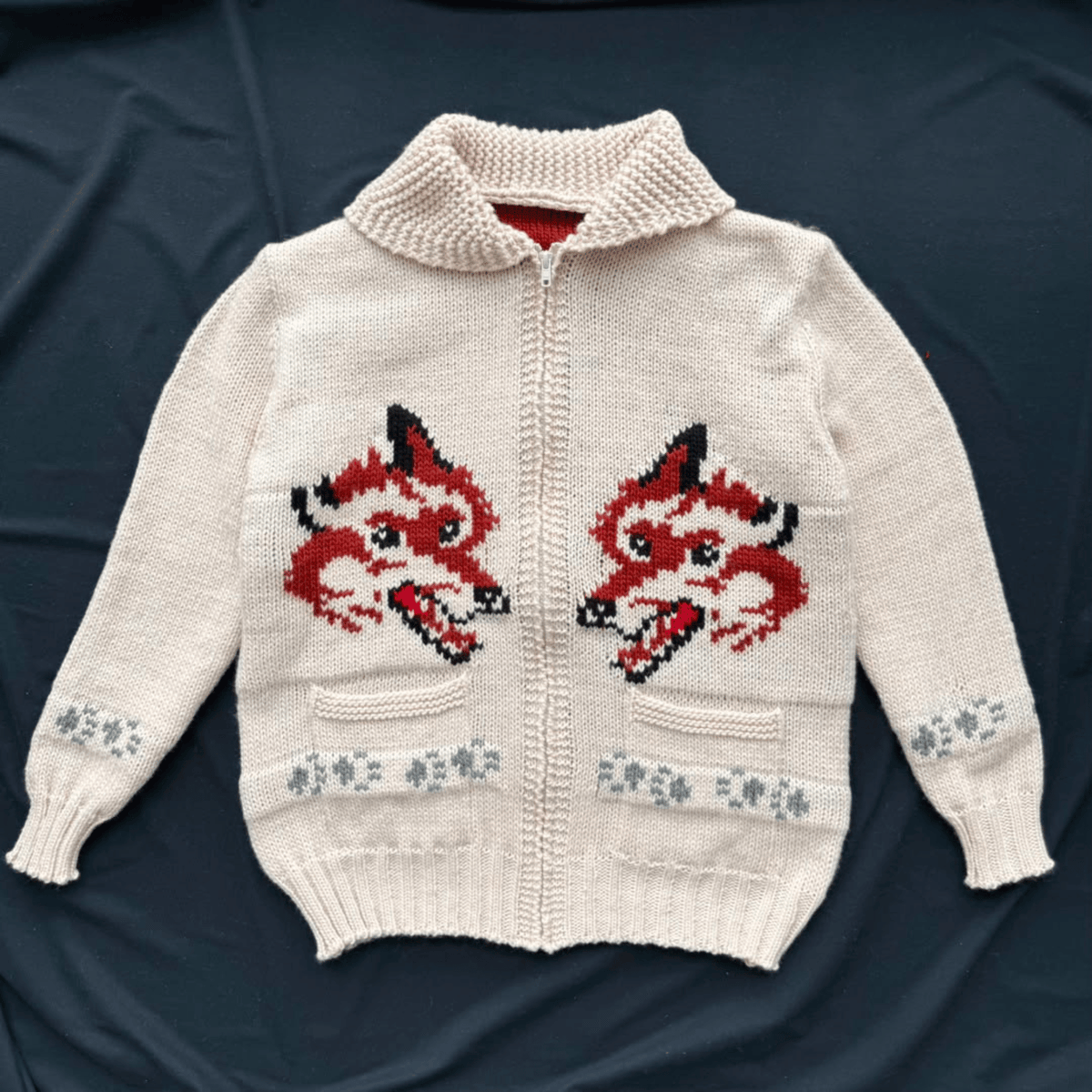An off-white knit zip-up cardigan sweater featuring two large red and black fox head motifs on the chest, with small gray floral patterns on the cuffs and hem, laid flat on a dark navy background.