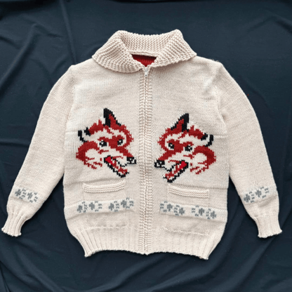 An off-white knit zip-up cardigan sweater featuring two large red and black fox head motifs on the chest, with small gray floral patterns on the cuffs and hem, laid flat on a dark navy background.