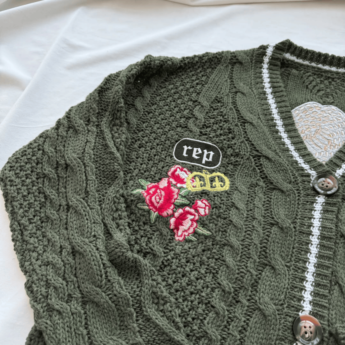An olive green cable-knit cardigan sweater laid flat on a white fabric background. The cardigan features white trim along the neckline and button placket, brown buttons, and colorful embroidered details on the chest, including a black 'rep' patch, yellow '44' text, and red roses.