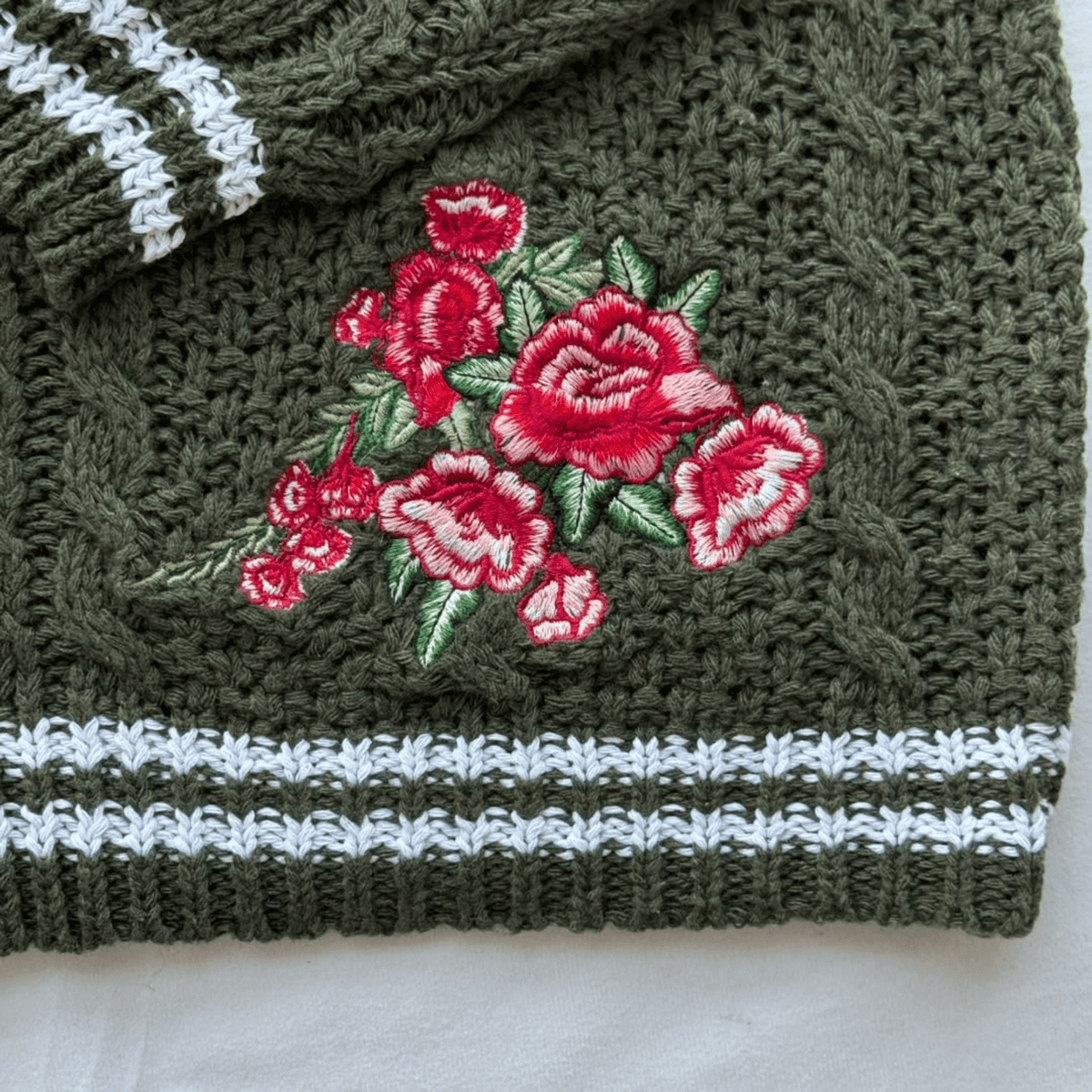 Close-up of a dark olive green knitted textile, likely a hat or scarf, featuring a red and pink floral embroidery with green leaves and white striped ribbing at the bottom.